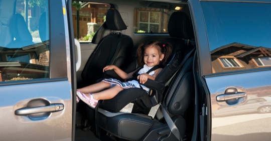a minivan with an open door, inside there is a little girl sitting in a buckled car seat and smiling