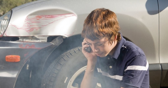 teenage boy sitting on the ground next to a silver car with a large dent and red scrapes, the teen is on the phone with a nervous look on his face