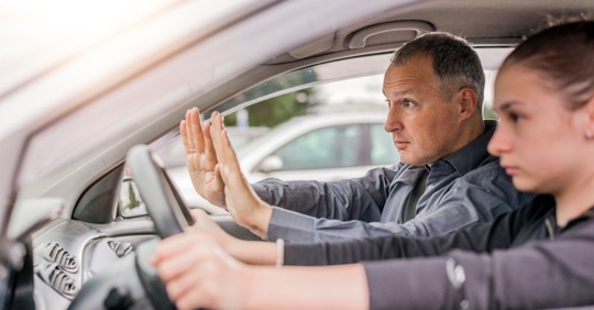 teenage girl and older man in a car, the girl is in the driver's seat and the man has a worried look on his face
